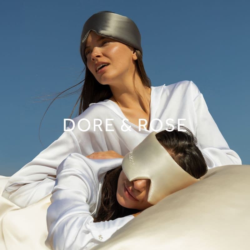 Dore & Rose: two women in white silk and silk eye masks against a blue sky, the Dore & Rose name across the picture